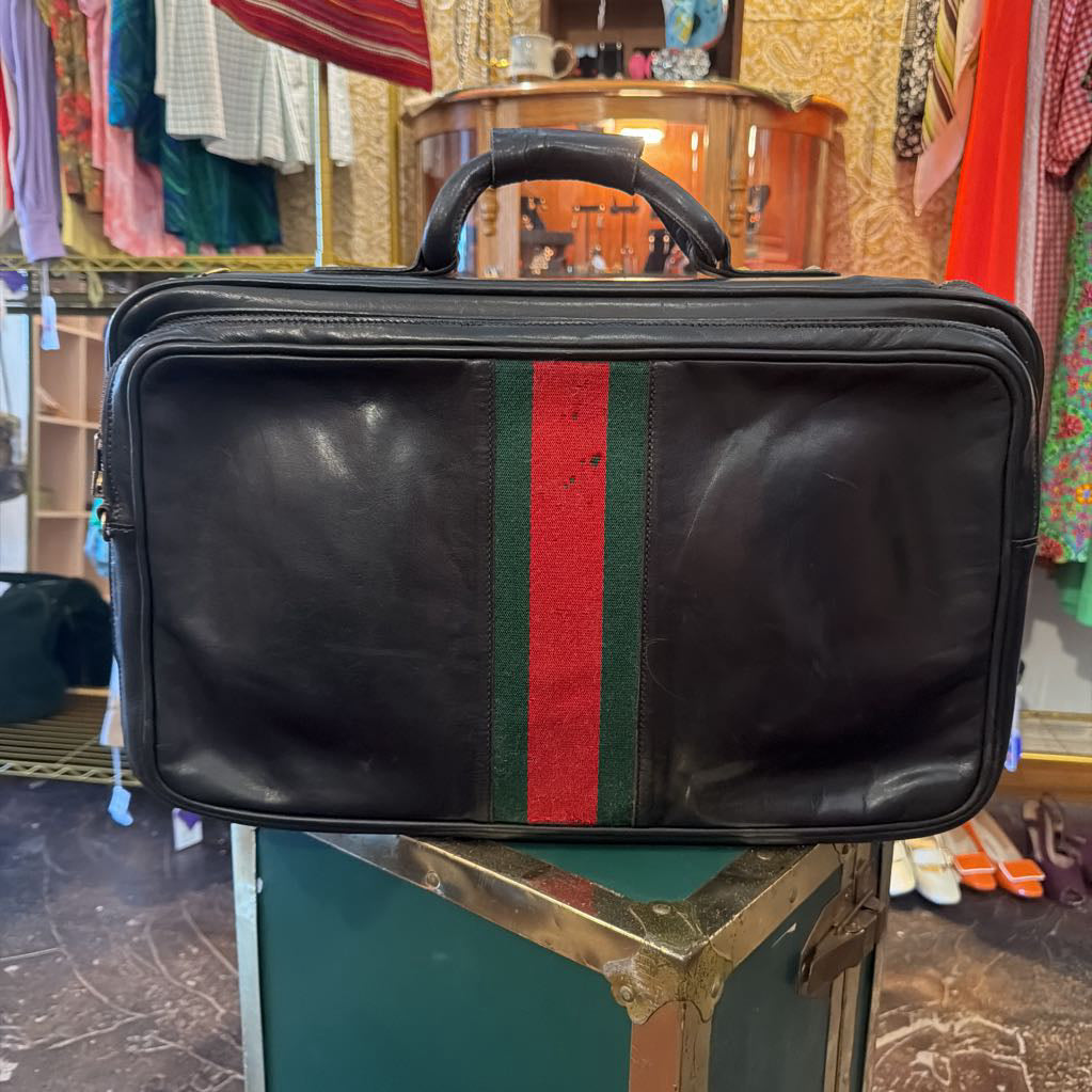 A black leather briefcase-style bag with a vertical green and red web stripe on the front, photographed in a retail or storage room setting on top of a green trunk.