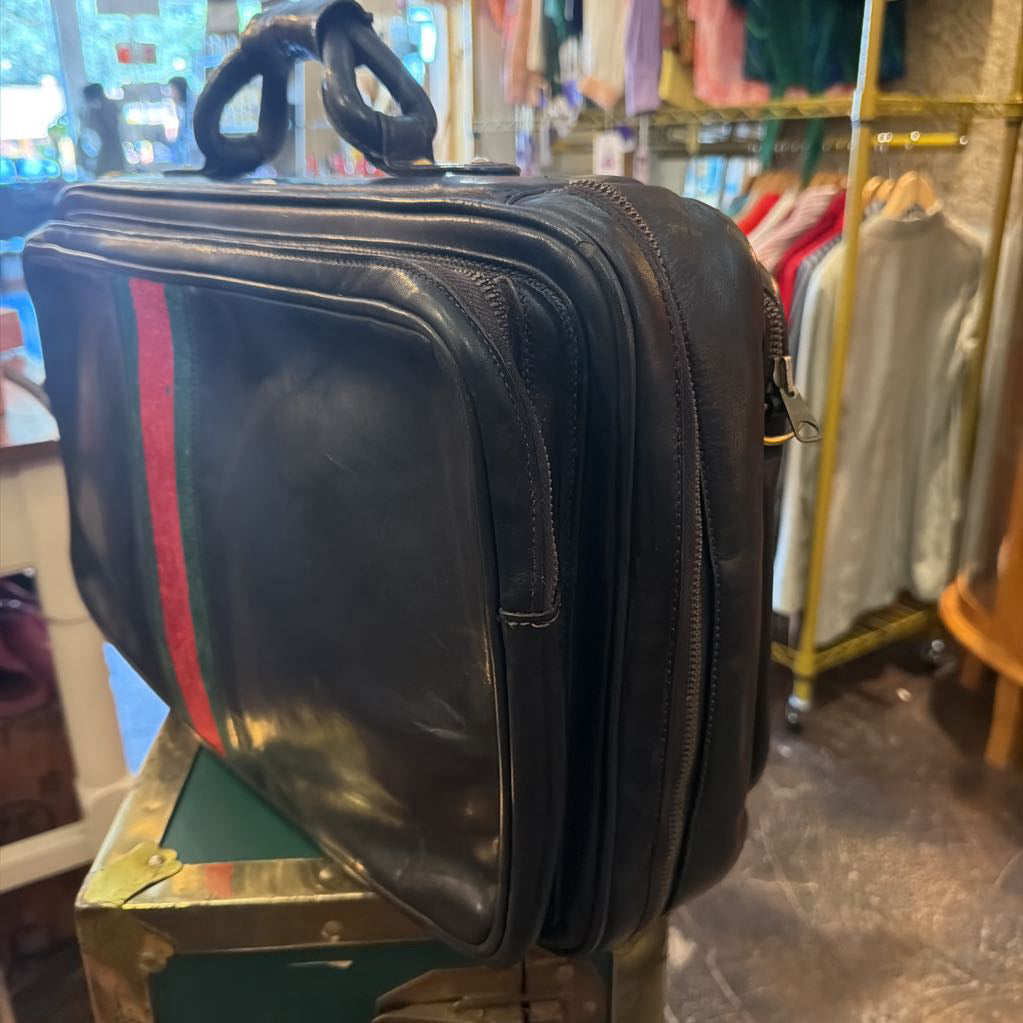 A black leather briefcase with the signature green and red web stripe on the side, resting on a vintage green and gold trunk inside a clothing store.