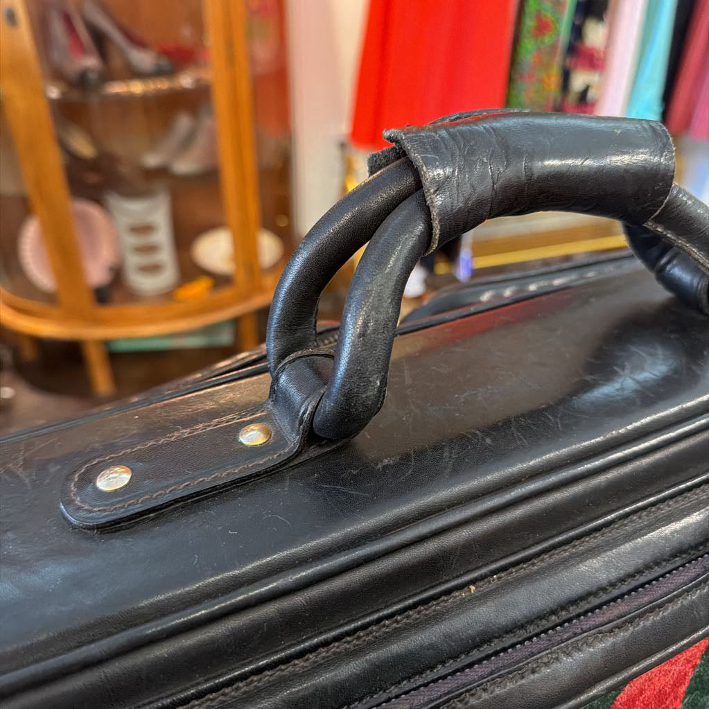 Close-up of a black leather suitcase handle with brass hardware, set against a blurred shop background.