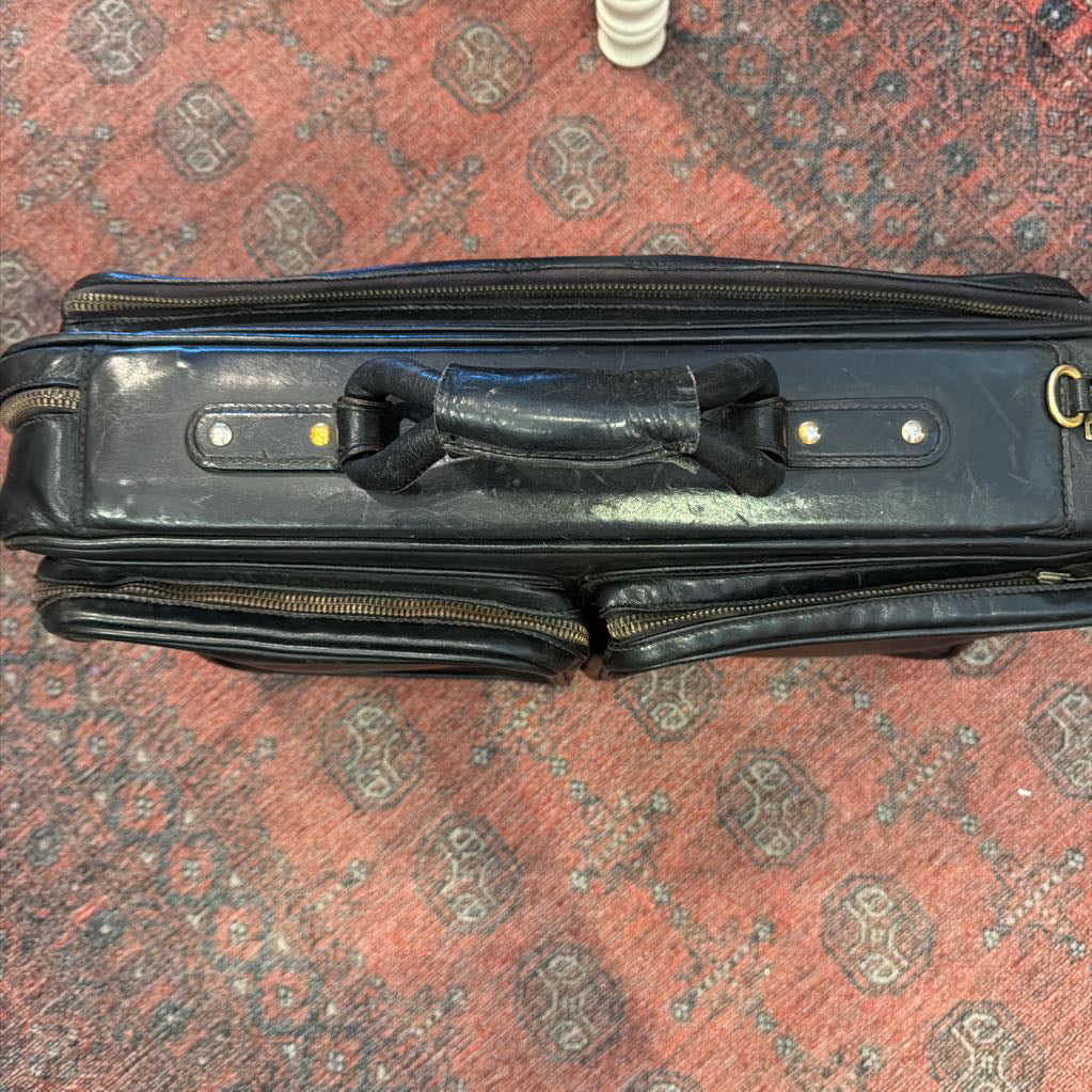 A black leather briefcase or laptop bag with a distressed handle, resting on a red patterned rug.