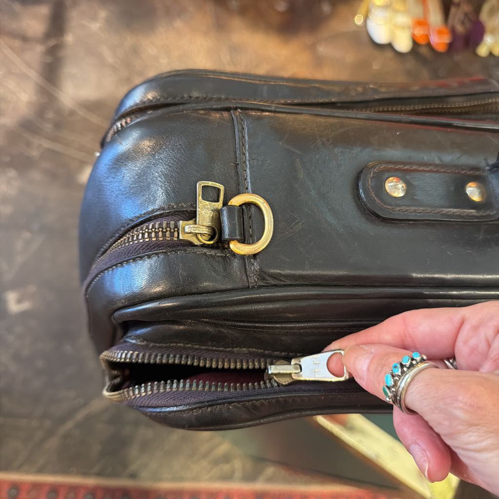 A close-up of a black leather bag with brass hardware, showing a hand pulling open a zipper. The hand has silver rings with turquoise accents.