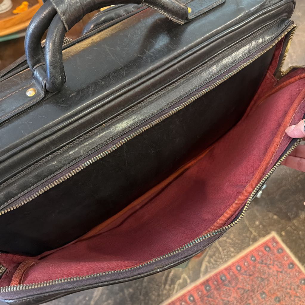 A close-up of an open black leather briefcase or satchel, showing a dark red or burgundy fabric interior lining. A hand is holding the zipper open.