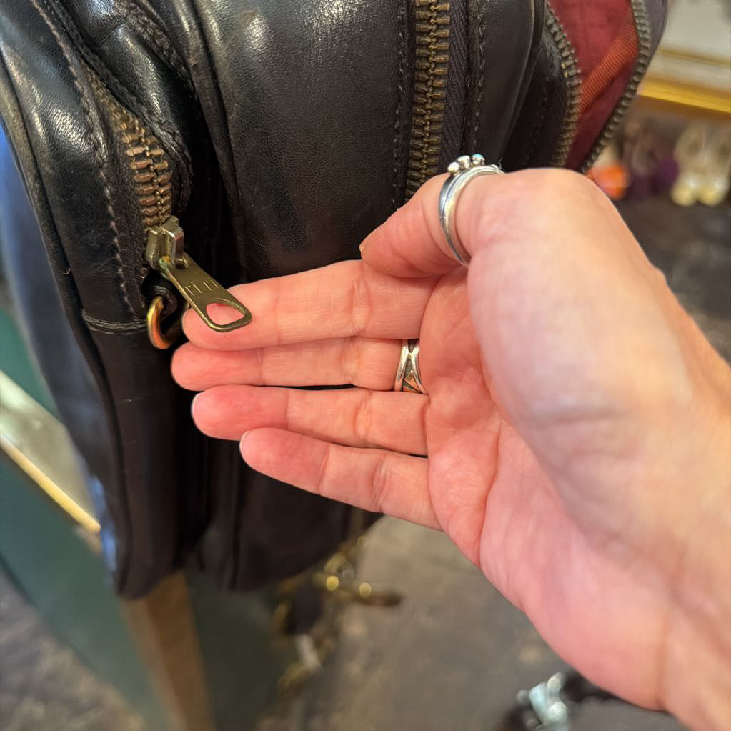 A close-up of a person's hand holding a brass zipper pull next to a black leather bag, with silver rings on the fingers.