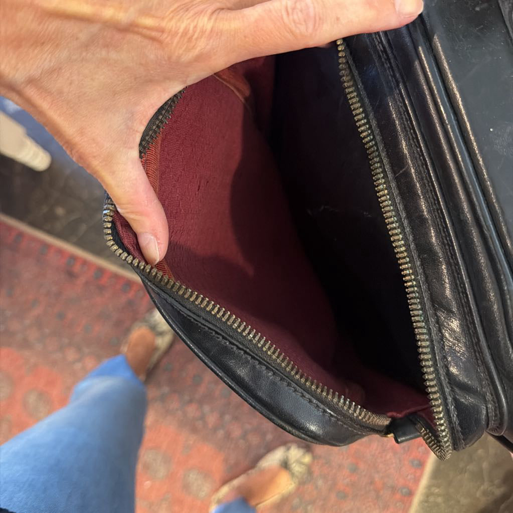 A close-up of a person's hand holding open a black leather zippered bag, showing a burgundy interior lining. The photo is taken from a top-down perspective on a patterned red carpet, with the person's blue jeans and bare feet visible at the bottom.