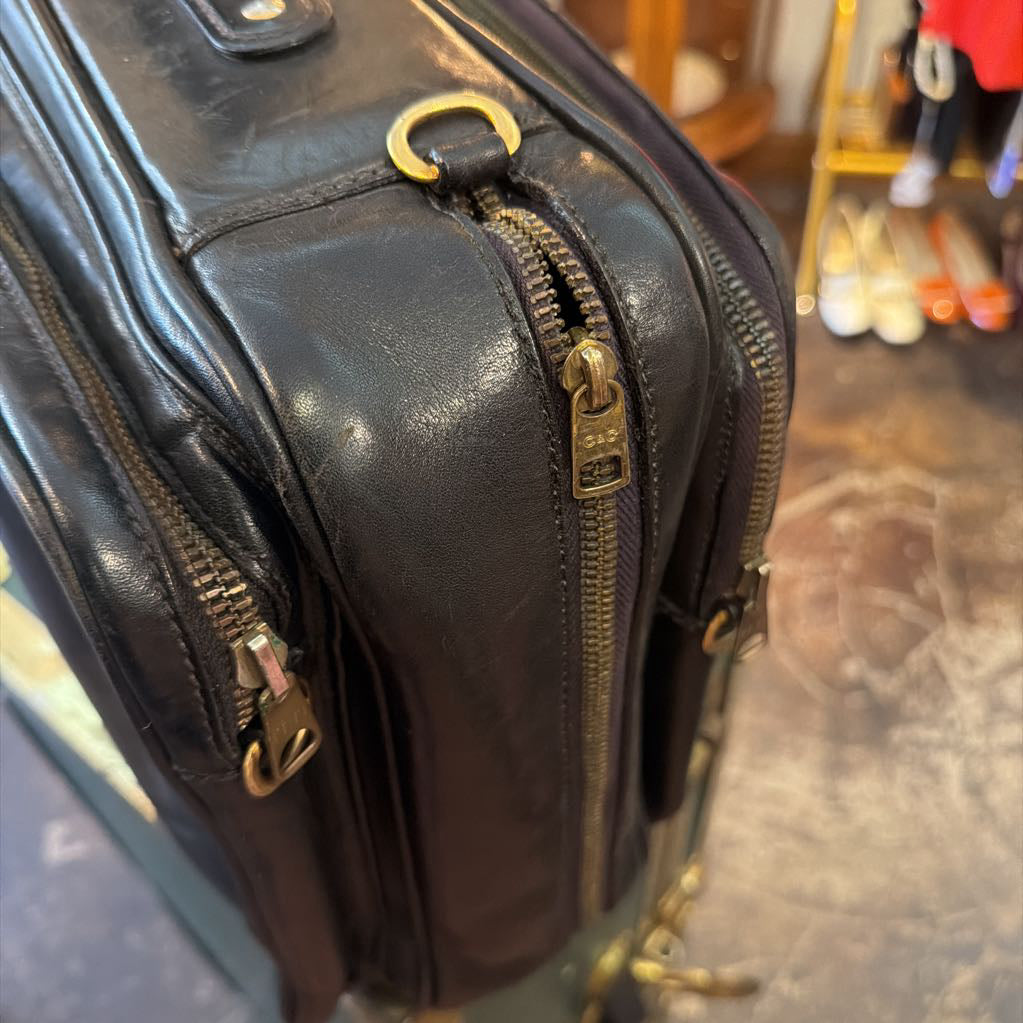 A close-up of a black leather suitcase corner showing dual brass zippers, a brass D-ring, and a worn leather surface, with a blurred store background.