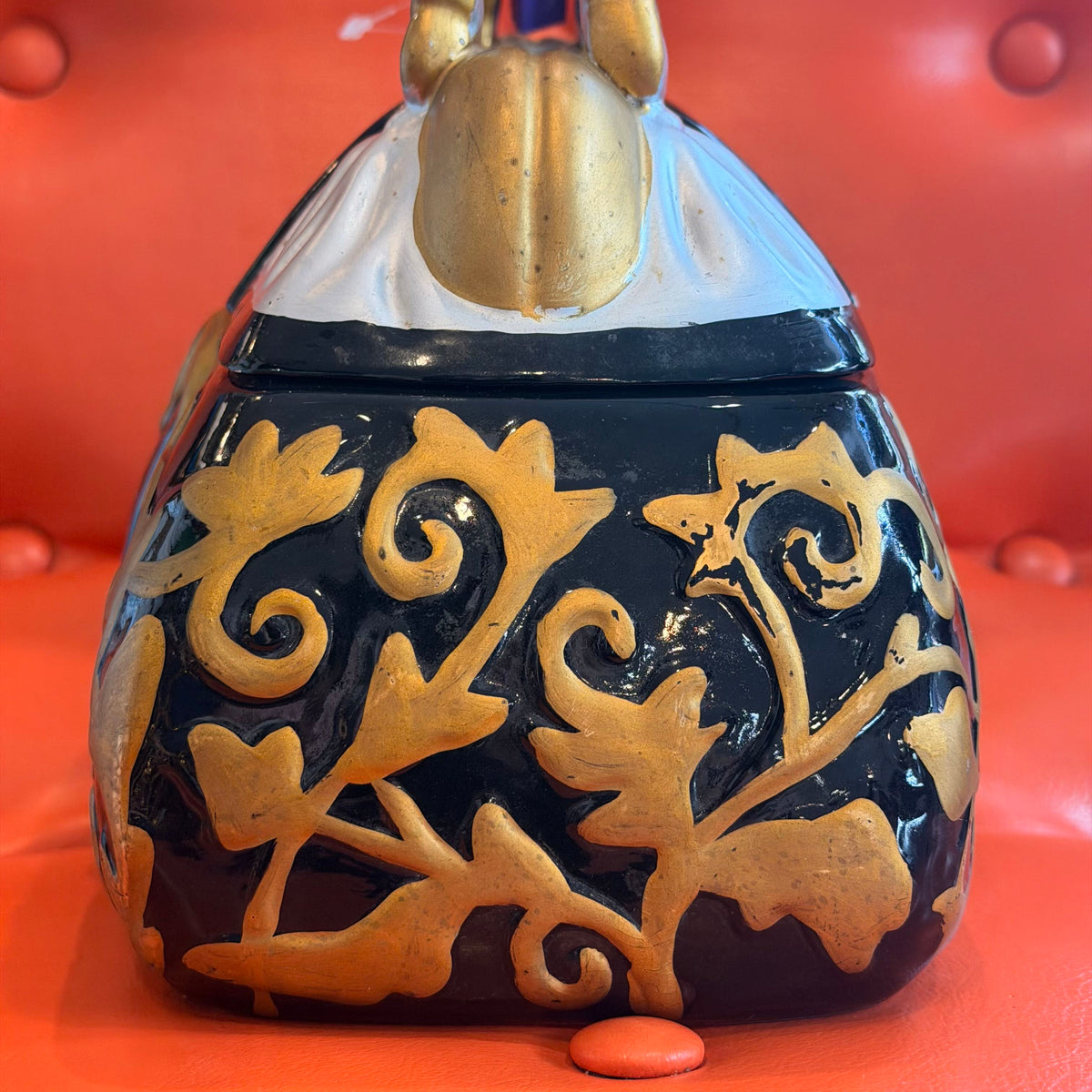 A decorative ceramic lidded container or candelabra-style vessel with a glossy black body, raised gold floral scroll pattern, and a sculpted gold top, photographed against a bright red tufted background.