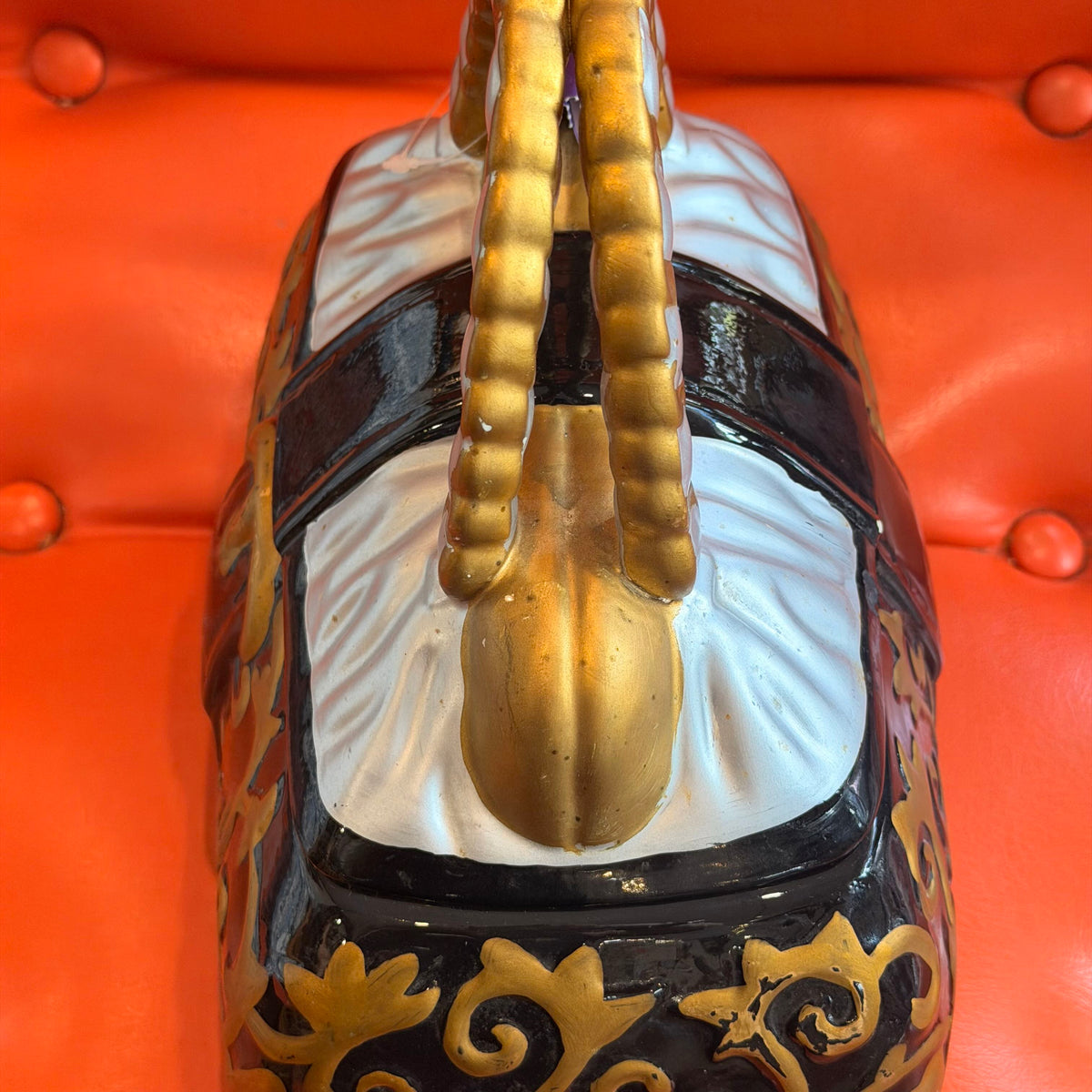 A close-up of an ornate handbag or decorative trunk with a glossy black body, white central panel, gold handles, and gold scrollwork, resting on a bright orange tufted surface.