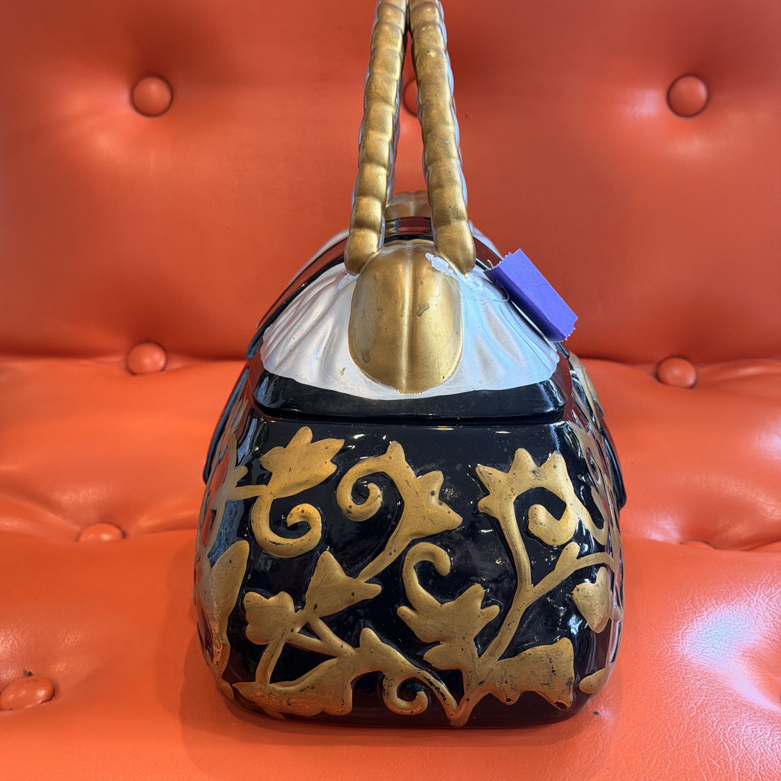 A vintage-style black handbag with ornate gold floral detailing, a white fabric top, and twisted gold handles, displayed on a bright orange tufted leather surface.
