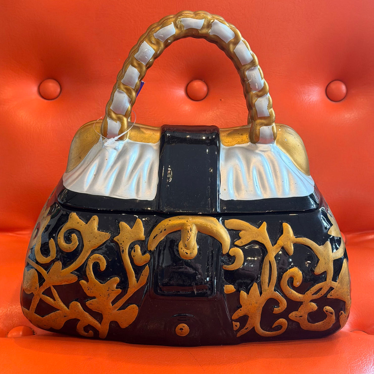 A decorative ceramic handbag-shaped object with a glossy black body, ornate gold scrollwork, white fabric-textured flap details, and a gold-and-white handle, set against a bright orange tufted background.