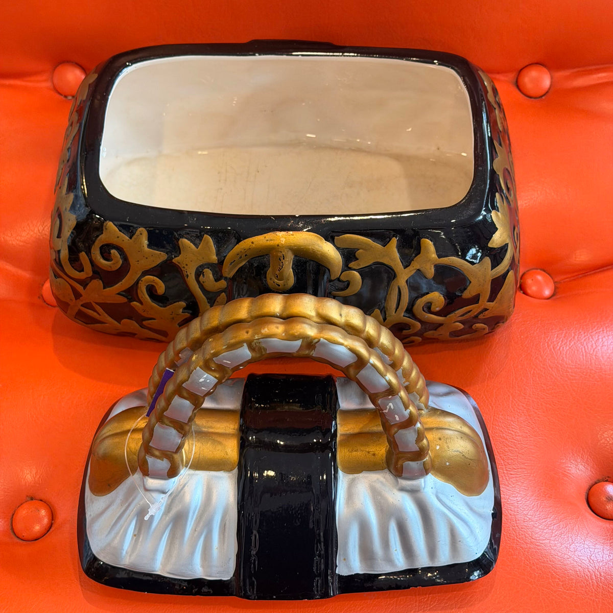 A decorative black and gold ornate box with a white interior, shown alongside a matching purse-shaped object on a bright orange tufted surface.
