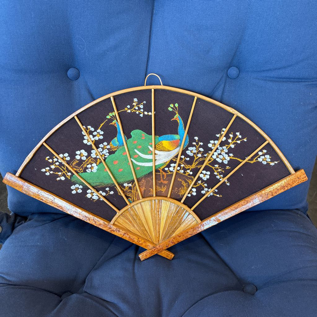 A black fabric and wooden fan wall decor with a painted peacock motif on a blue cushioned surface.