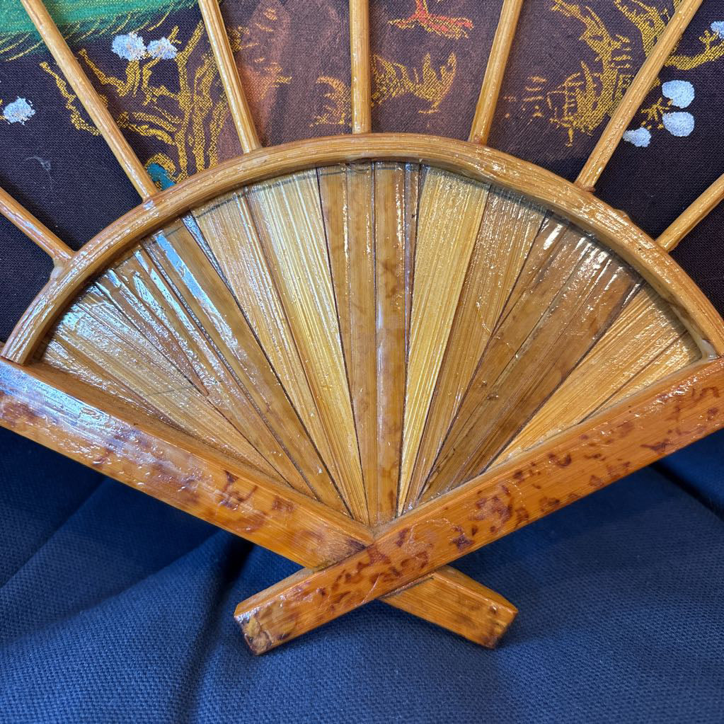 A black fabric and wooden fan wall decor with a painted peacock motif on a blue cushioned surface.