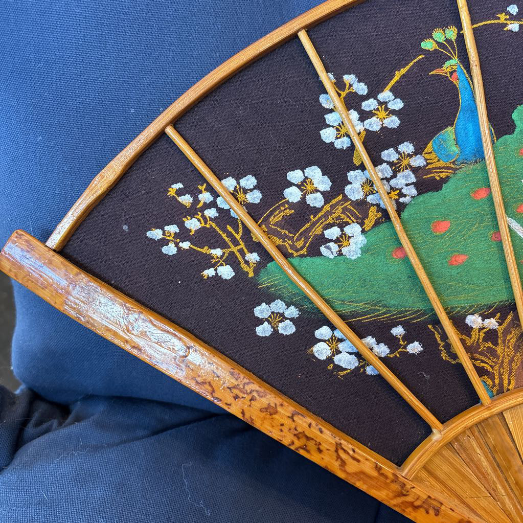 A black fabric and wooden fan wall decor with a painted peacock motif on a blue cushioned surface.