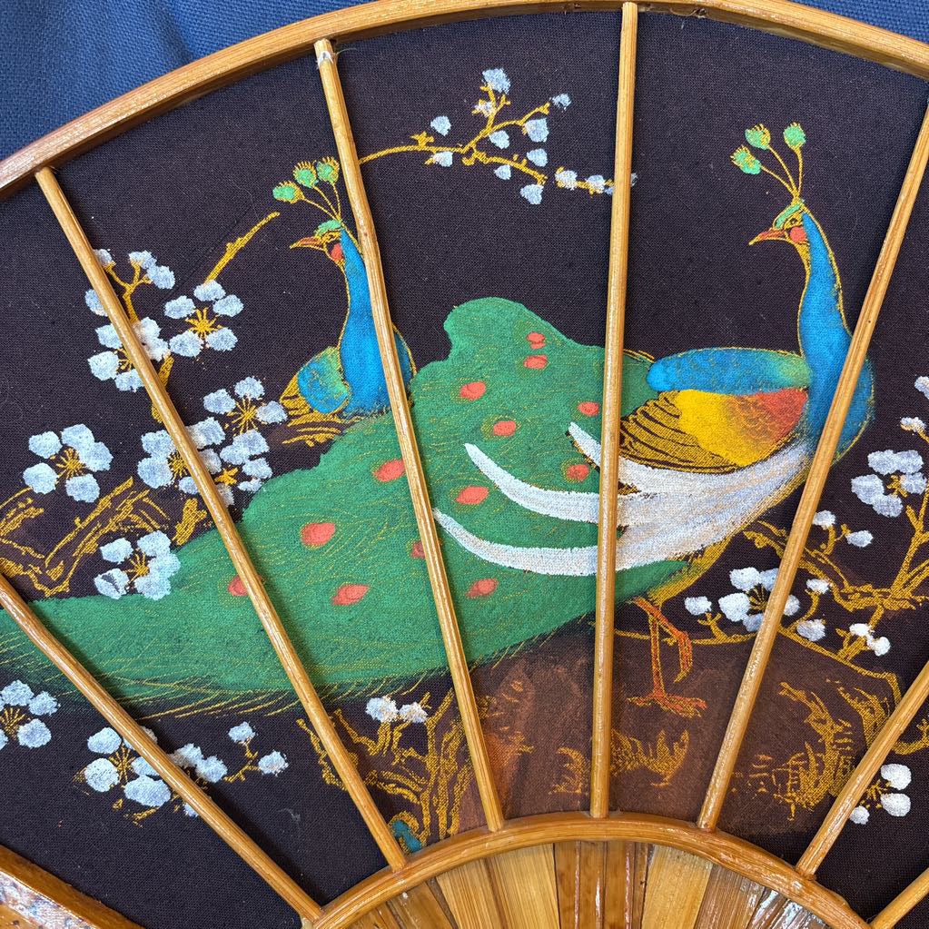 A black fabric and wooden fan wall decor with a painted peacock motif on a blue cushioned surface.