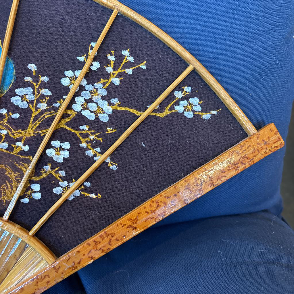 A black fabric and wooden fan wall decor with a painted peacock motif on a blue cushioned surface.