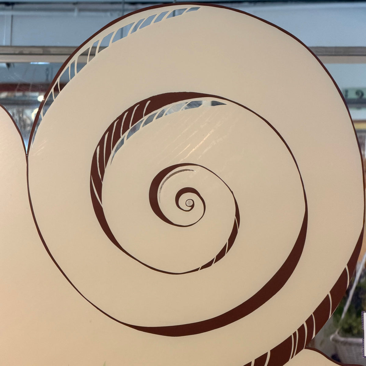 A close-up of a large spiral-shaped decorative sign or wall art piece with a cream background and dark brown linework, photographed indoors with some blurred background elements visible.