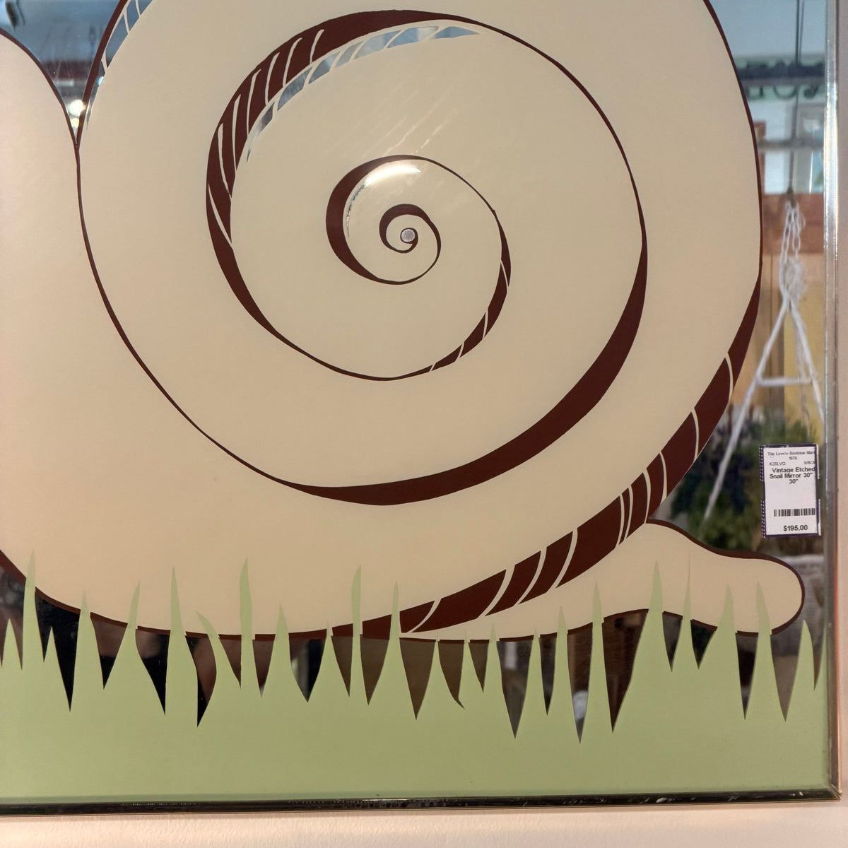 A decorative wall art print or panel featuring a large stylized snail shell in cream and dark brown above a pale green grassy base, photographed close up with some reflections and a small price tag visible on the right side.
