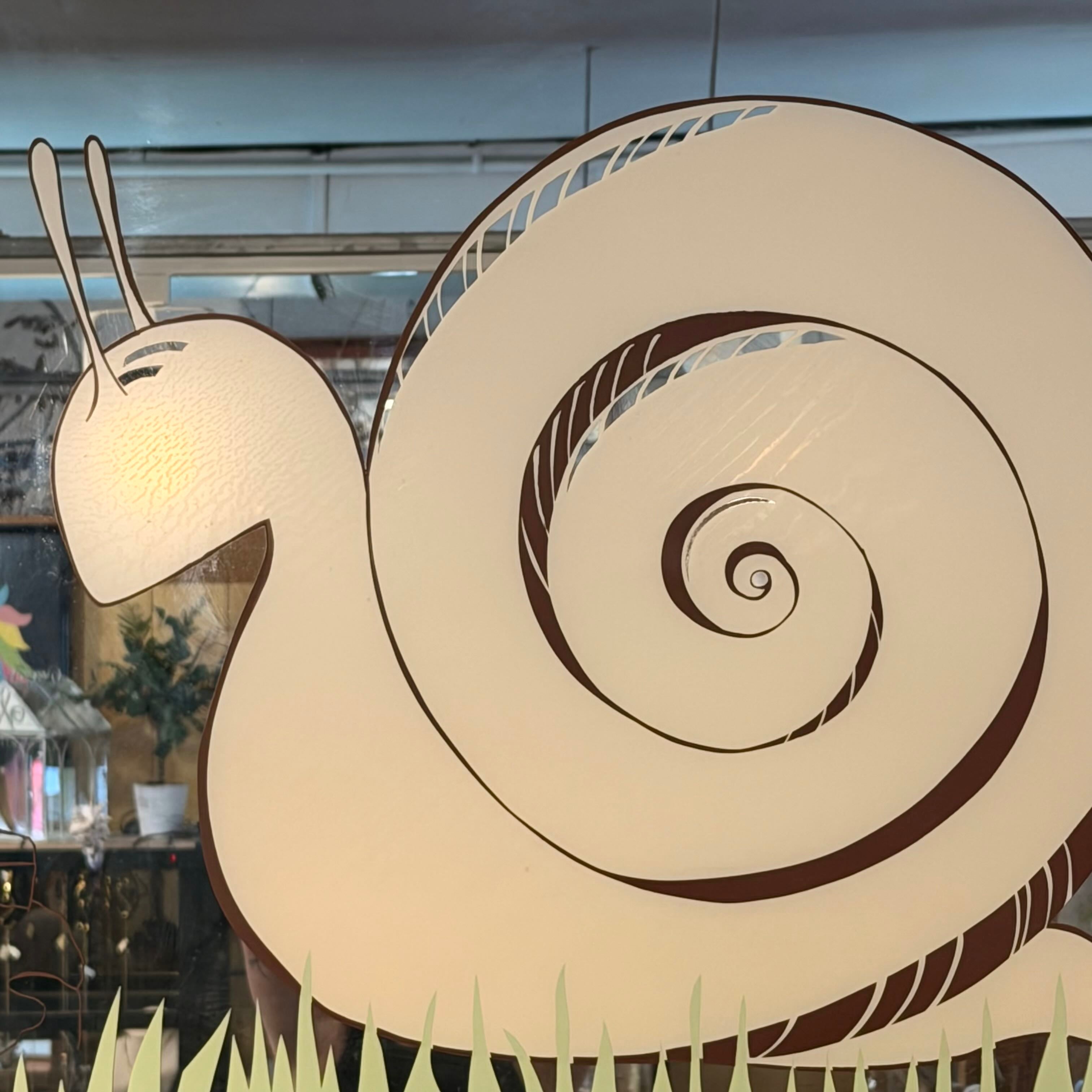 A large decorative snail cutout or wall decor piece with a cream-colored body and shell outlined in dark brown, shown indoors against a window with reflections and store items visible in the background.