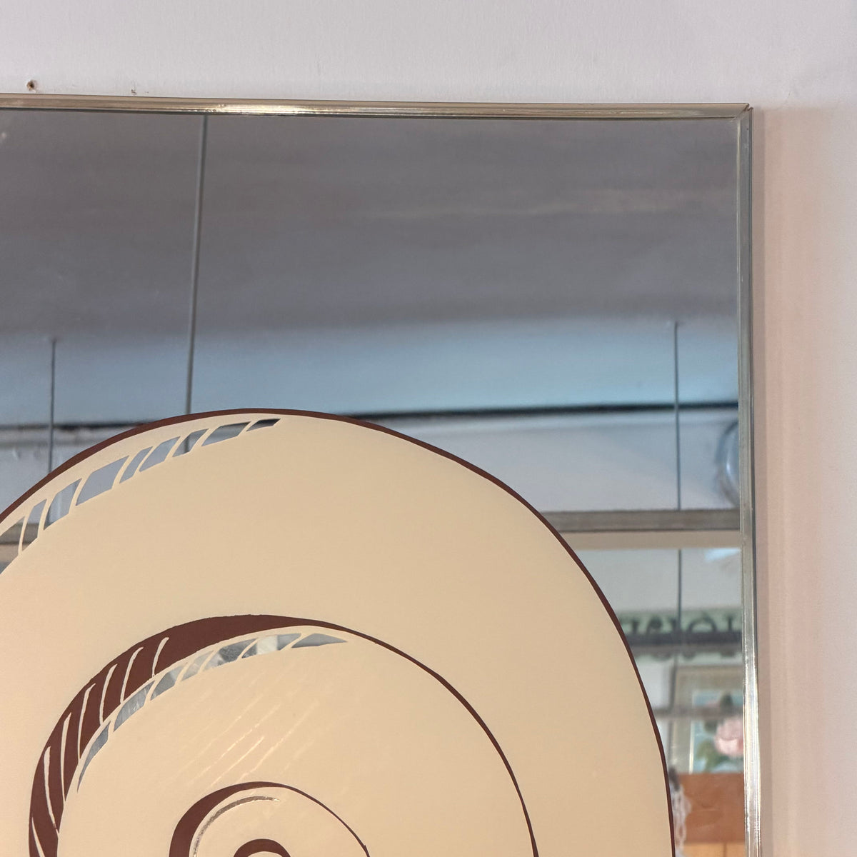 A close-up of a decorative circular wall mirror with a cream and brown spiral pattern, reflecting a room interior with a window and ceiling.