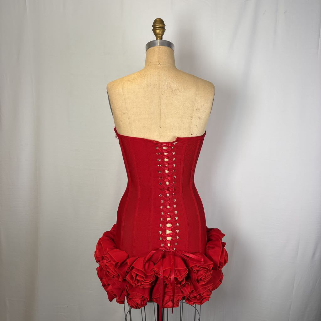 A red strapless corset-style dress displayed on a fabric dress form, viewed from the back. The dress has lace-up back detailing and a ruffled floral skirt.
