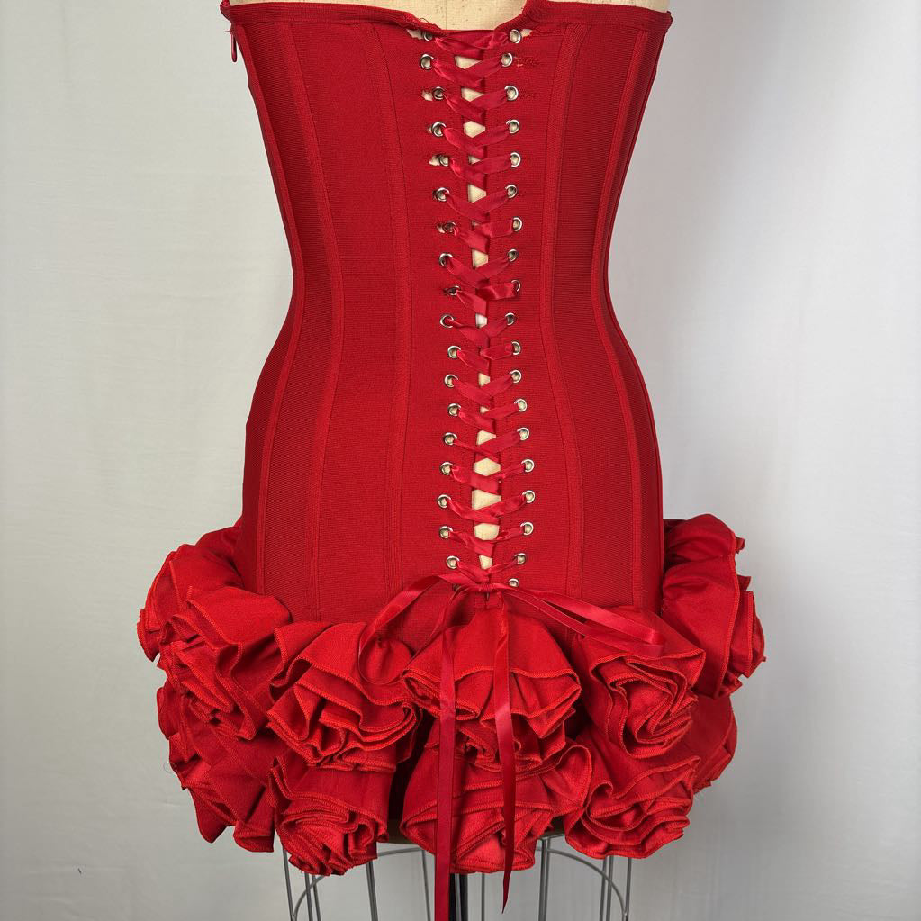 A bright red corset-style top or dress bodice displayed on a mannequin, featuring a lace-up front with silver eyelets and a dramatic ruffled skirt at the bottom.