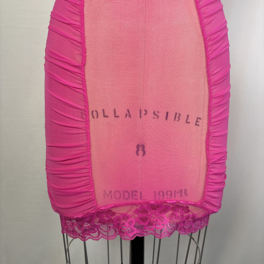 A bright pink sheer mesh garment or fabric panel with gathered ruffled trim on the sides and a lace hem, displayed on a mannequin torso against a plain light background. Faint text on the center reads 'COLLAPSIBLE' and 'MODEL 1991-11'.
