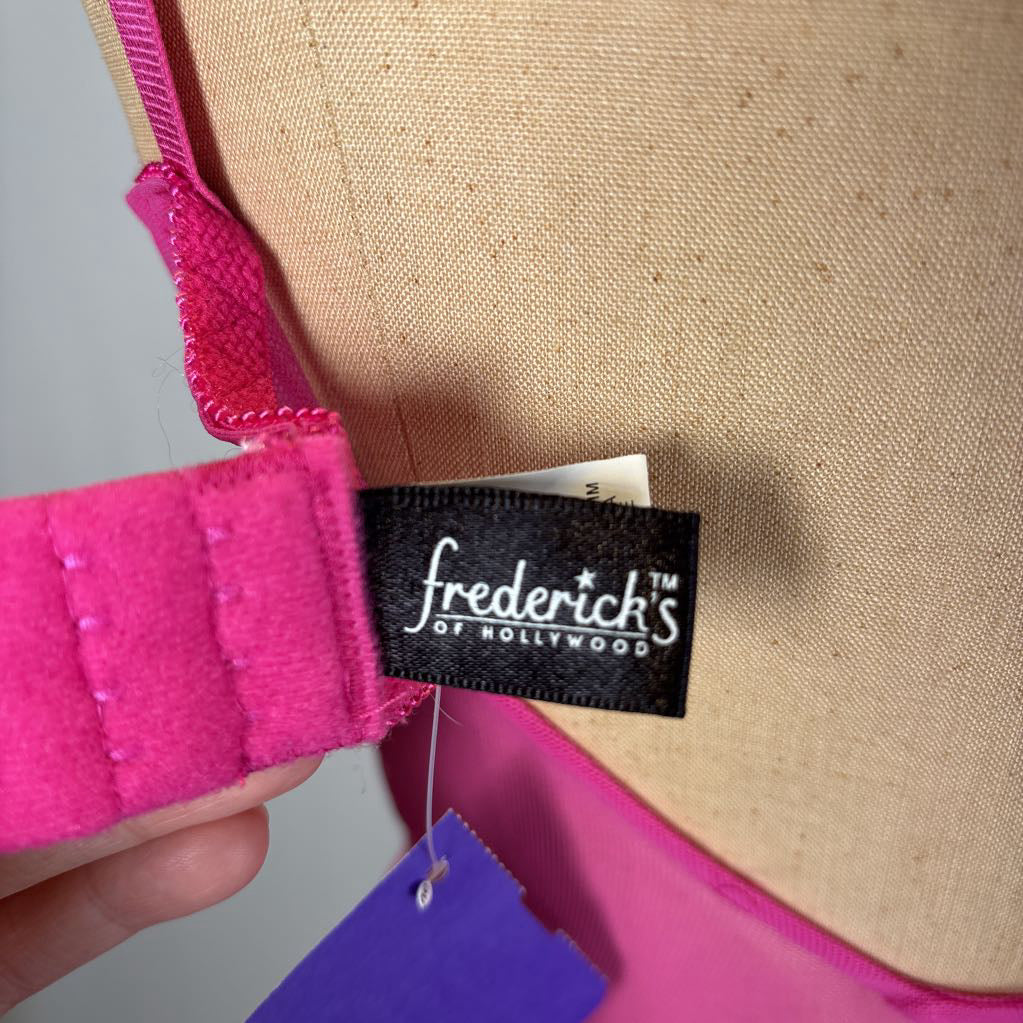 Close-up of a bright pink bra strap attached to a black fabric brand label reading 'frederick's OF HOLLYWOOD', with part of a beige bra cup and a purple hangtag visible.