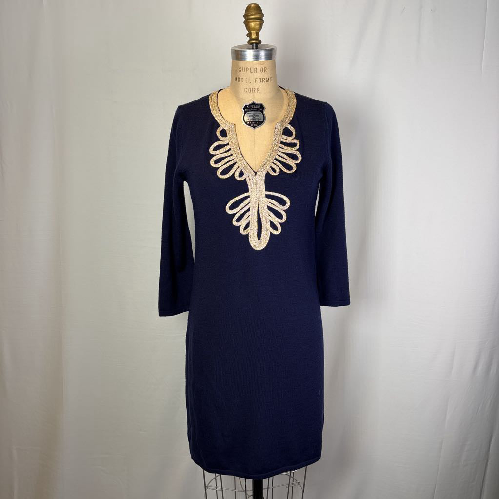 A navy blue knit dress displayed on a dress form mannequin against a white fabric backdrop. The dress features a V-neckline embellished with a large, decorative beige crochet or lace overlay on the chest and shoulders, and three-quarter sleeves.