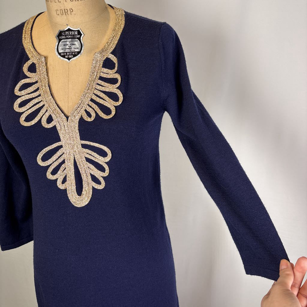 A navy blue long-sleeve knit top displayed on a tan dress form mannequin, featuring a decorative beige embroidered V-neckline pattern. A hand is holding the sleeve at the right edge against a plain light background.
