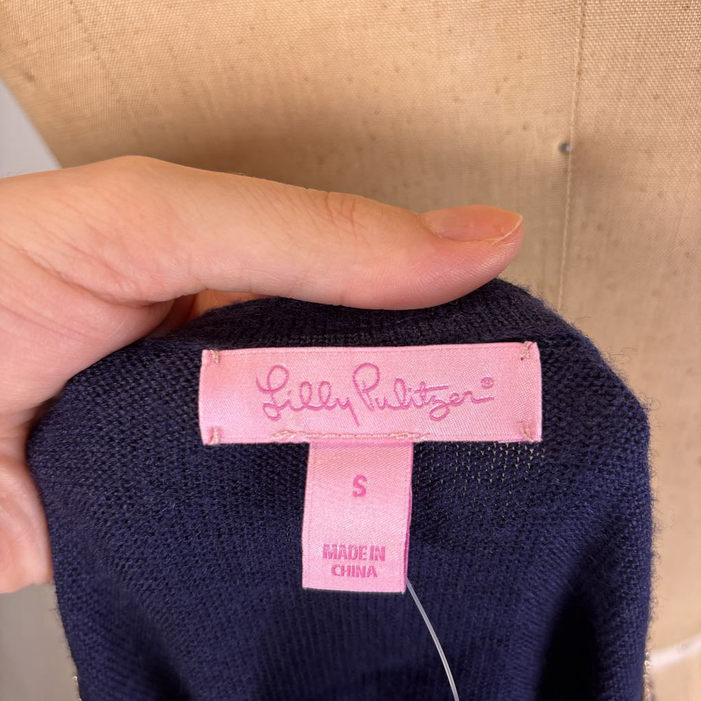 A close-up of a hand holding a navy blue knit garment, showing a pink sewn-in label that reads 'Lilly Pulitzer' and 'S MADE IN CHINA'.