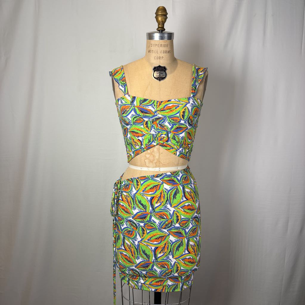 A two-piece matching set consisting of a sleeveless crop top and a high-waisted mini skirt, both featuring a vibrant multicolor abstract pattern in green, orange, yellow, and blue on a white background. The set is displayed on a vintage wooden dress form mannequin against a neutral gray fabric backdrop.