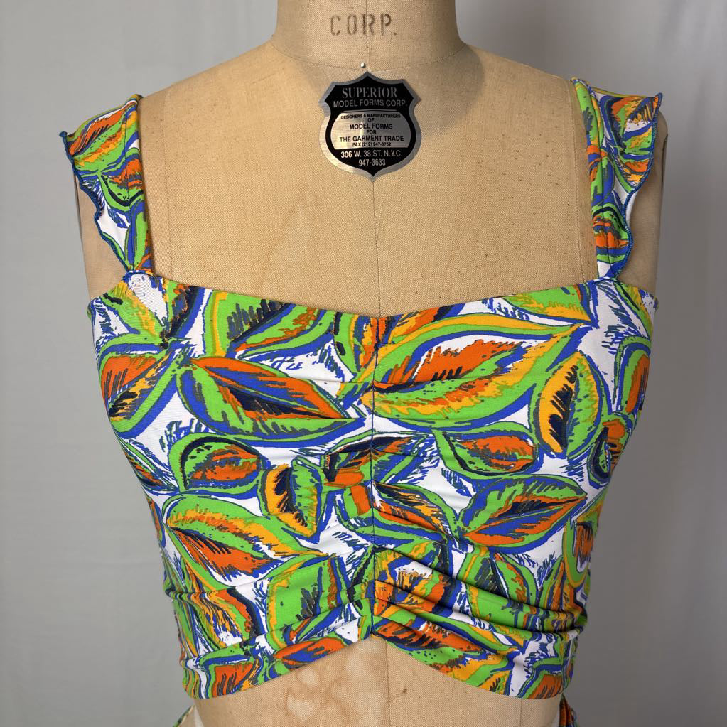 A sleeveless cropped top with a colorful tropical leaf print, displayed on a tan burlap mannequin torso. The print features green, orange, blue, and white leaves.