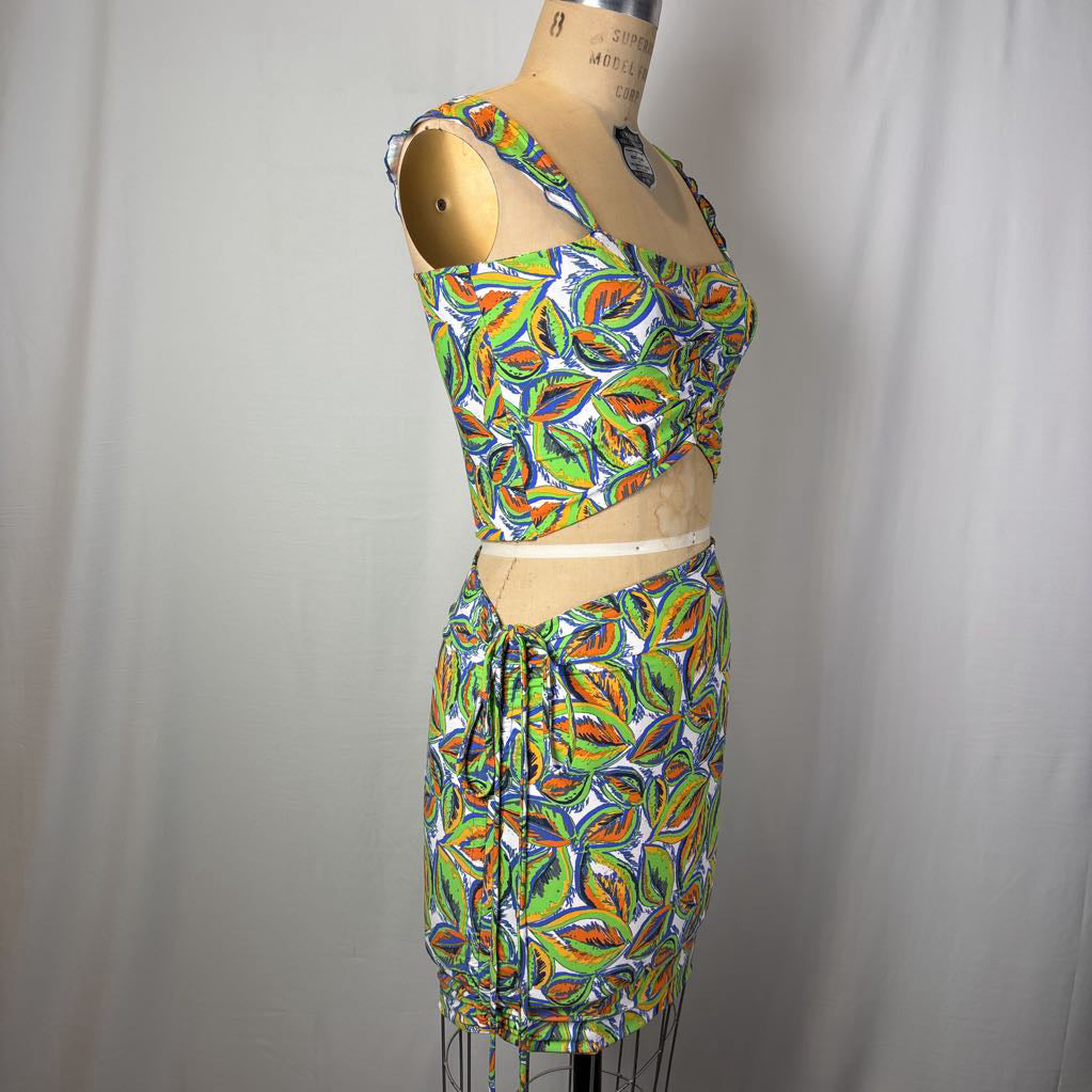 A two-piece matching set consisting of a sleeveless crop top and a high-waisted mini skirt, both featuring a vibrant multicolor abstract pattern in green, orange, yellow, and blue on a white background. The set is displayed on a wooden dress form mannequin against a light gray fabric backdrop.