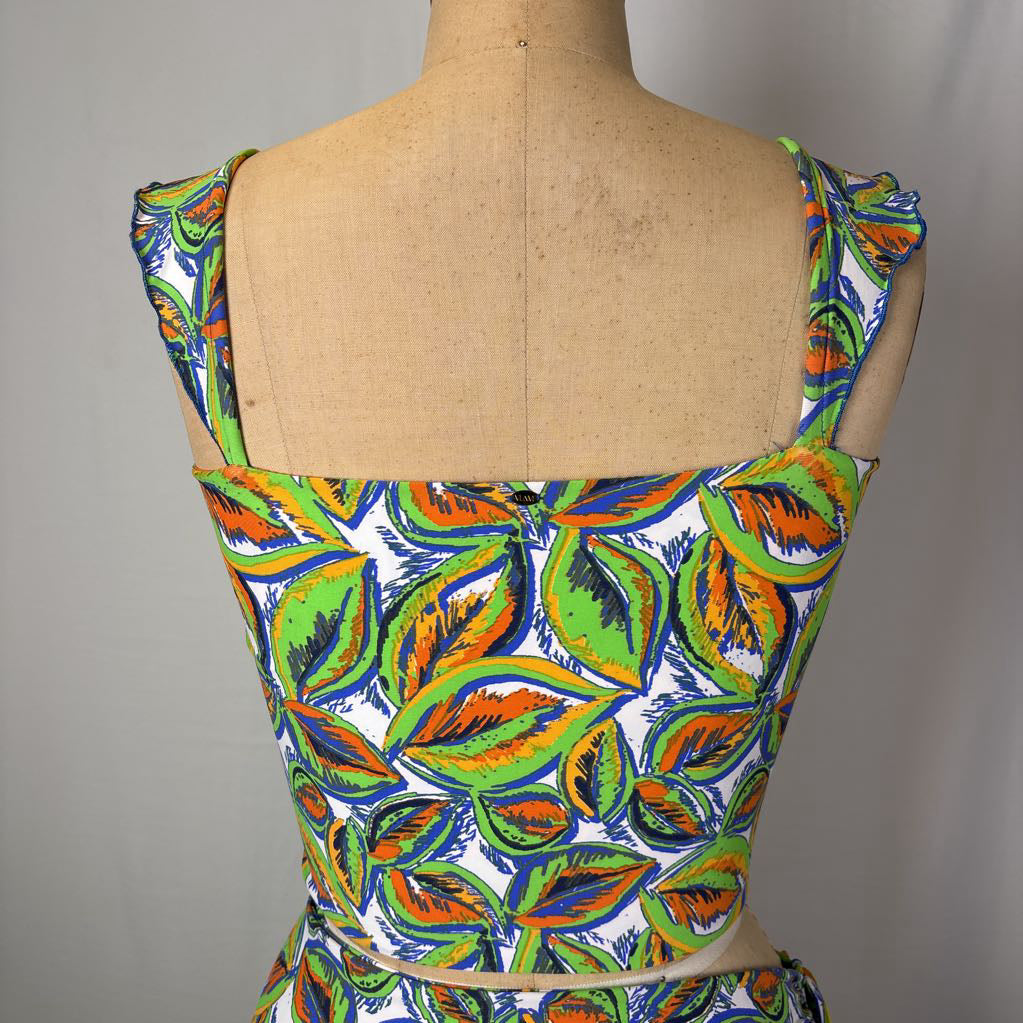 A sleeveless dress or bodice displayed on a tan dress form, featuring a bold leaf print in green, orange, blue, and white on a light gray background.