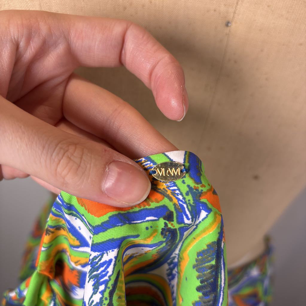 A close-up of a hand holding a colorful patterned fabric piece with a small gold 'MIMI' brand tag, positioned in front of a beige dress form.