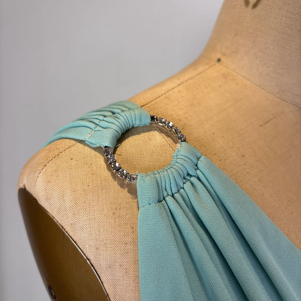 A close-up of a sage green draped fabric accessory with a rhinestone-embellished circular ring, displayed on a tan fabric dress form.