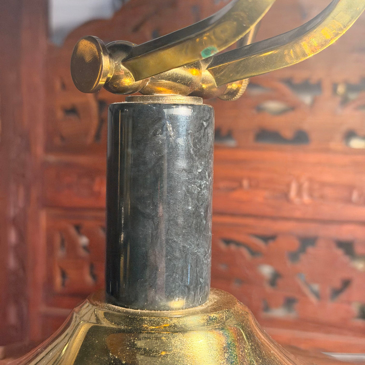 Close-up of a brass table lamp base with a polished black marble or dark stone cylindrical collar, set against a blurred reddish-brown carved wooden background.