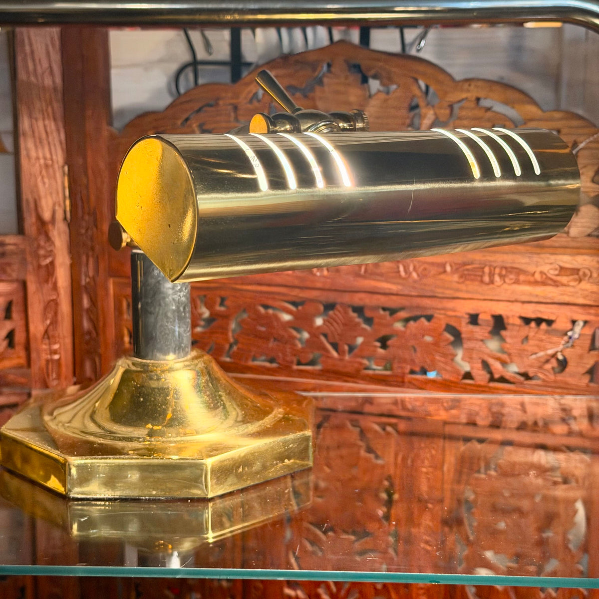 A polished brass desk lamp with a cylindrical shade, mounted on a hexagonal base, displayed on a glass shelf in front of a carved wooden screen.