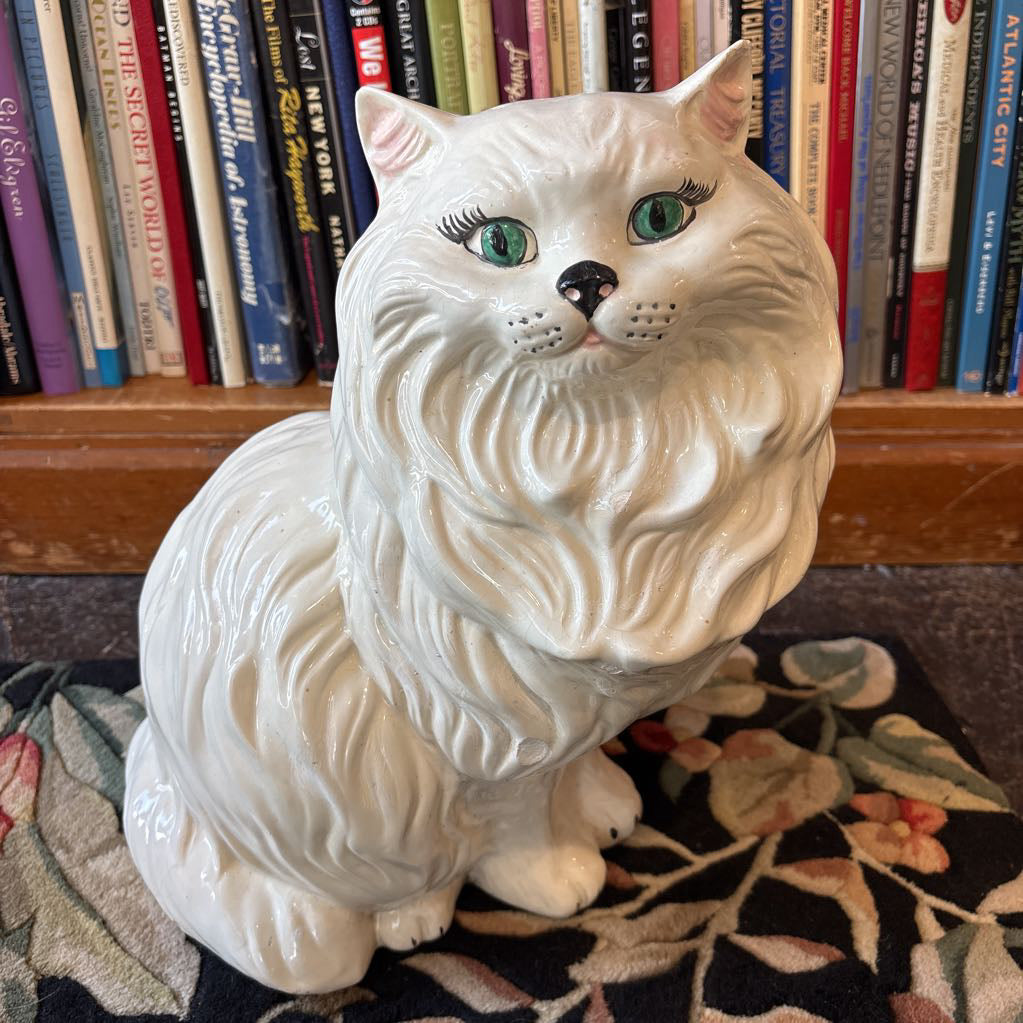A large white ceramic cat figure with green eyes and long eyelashes sitting on a floral rug against a bookcase. 