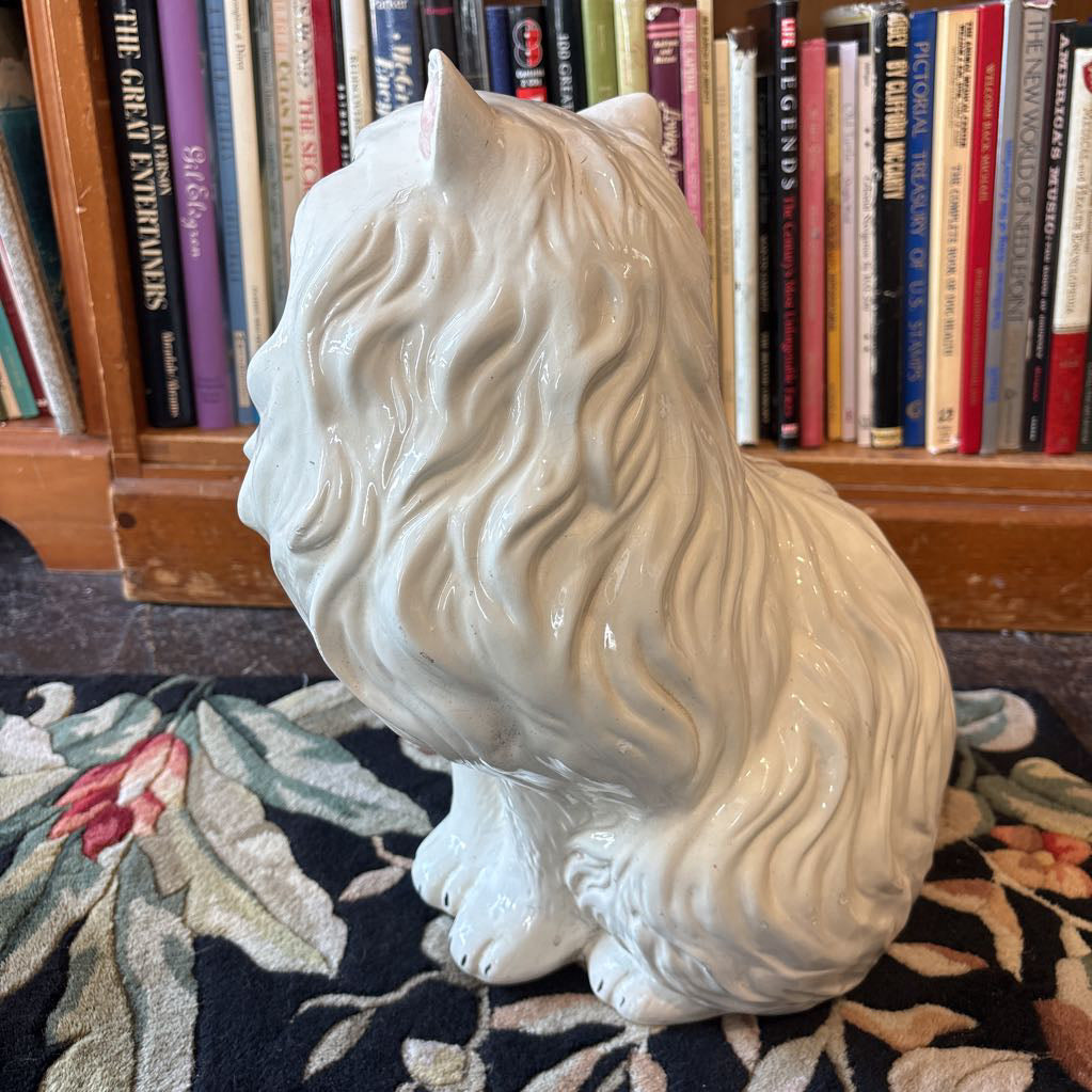 A large white ceramic cat figure with green eyes and long eyelashes sitting on a floral rug against a bookcase. 