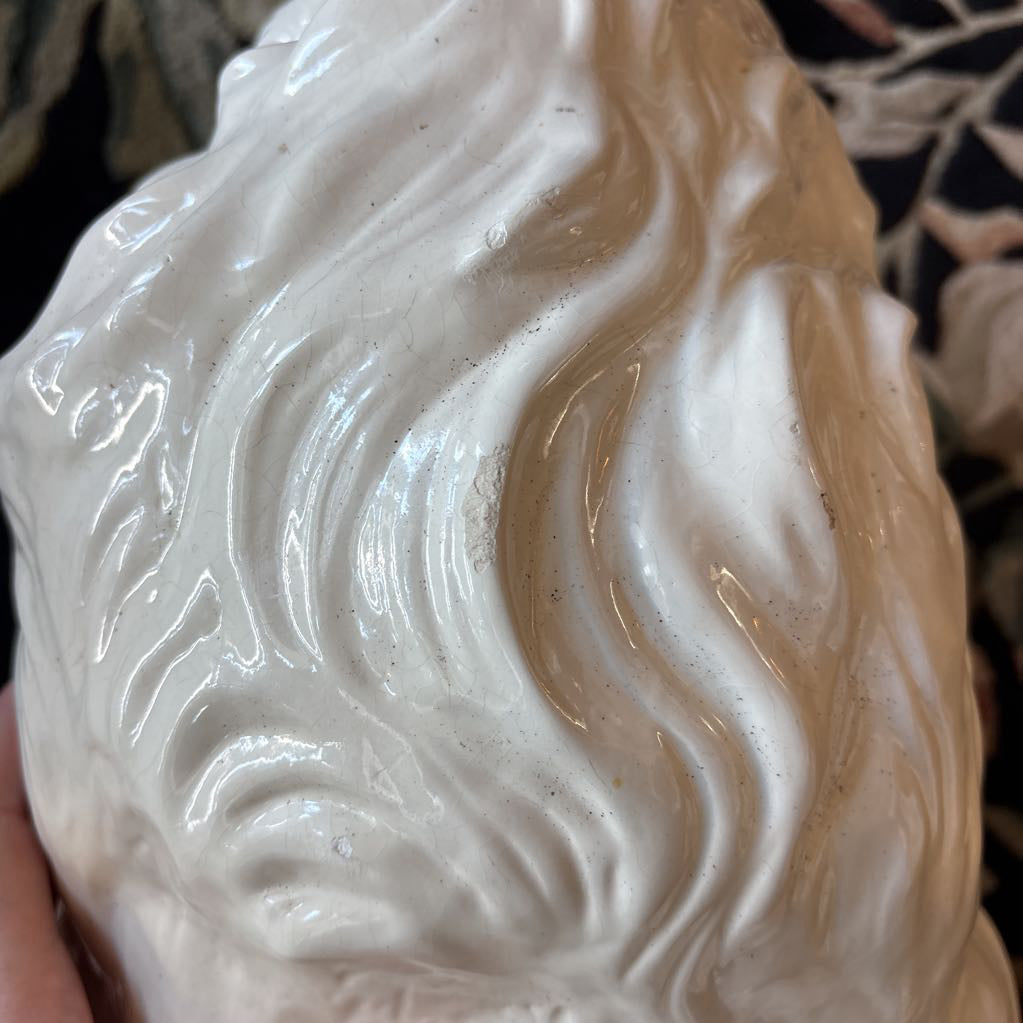 A close-up of a glossy white sculptural object with flowing, wave-like ridges, held against a dark background with some blurred decorative elements visible.