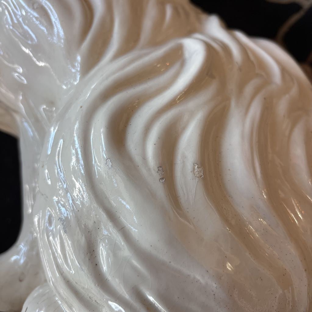 A close-up of a glossy white sculptural object with flowing, wave-like ridges, held against a dark background with some blurred decorative elements visible.