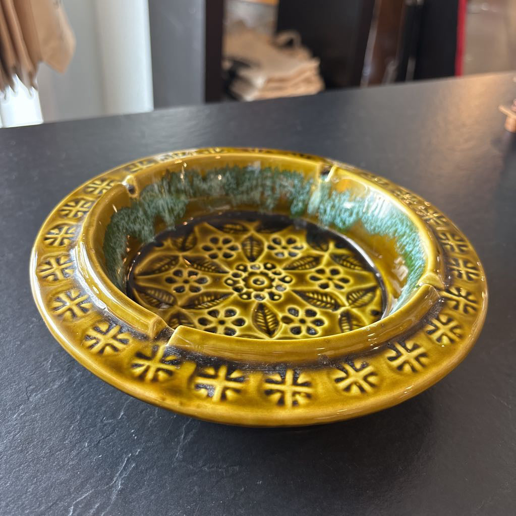 A round ceramic ashtray with a textured yellow-green glaze and embossed floral patterns, resting on a dark tabletop.