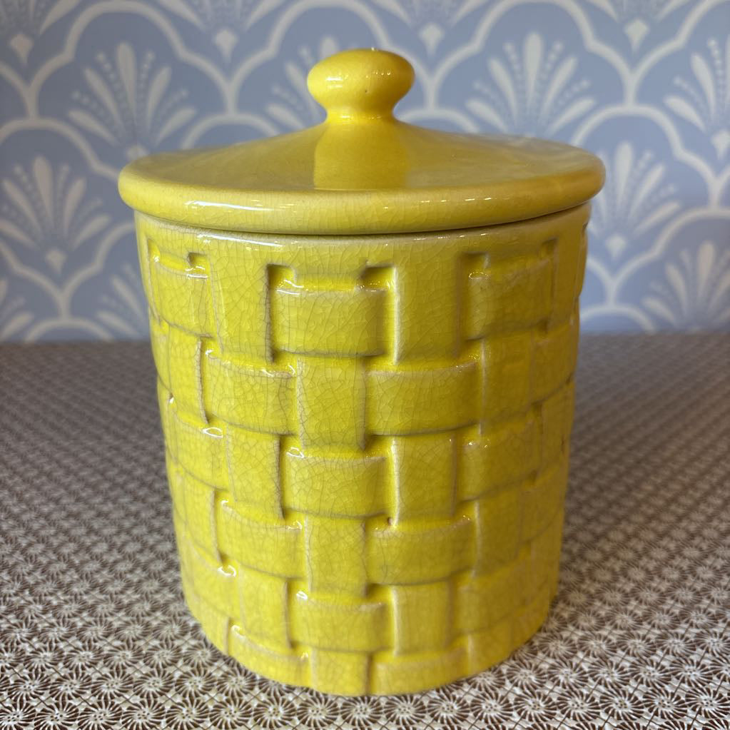 A yellow basket weave textured ceramic canister with lid on a wallpapered surface. 