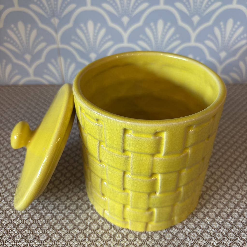 A yellow basket weave textured ceramic canister with lid on a wallpapered surface. 