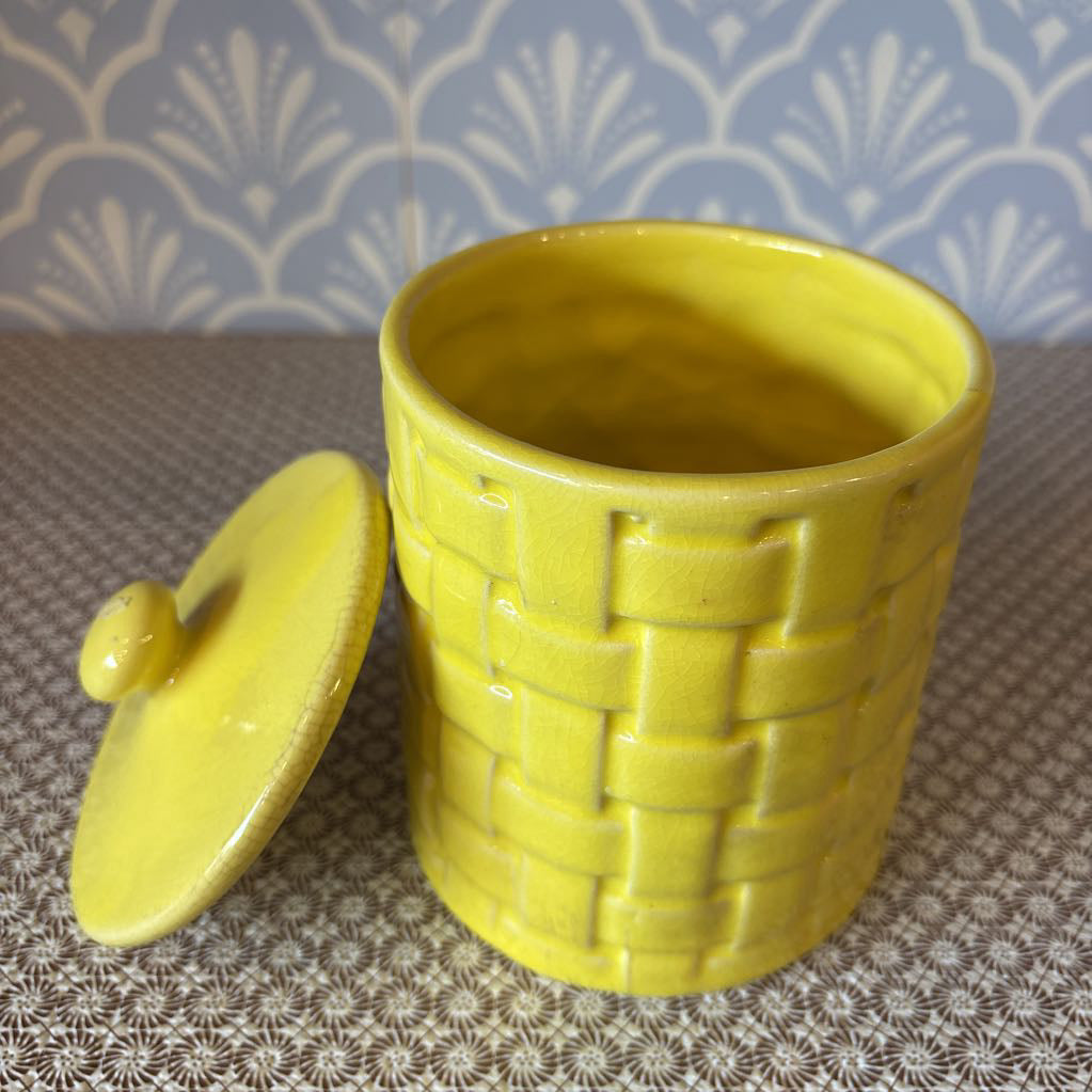 A yellow basket weave textured ceramic canister with lid on a wallpapered surface. 