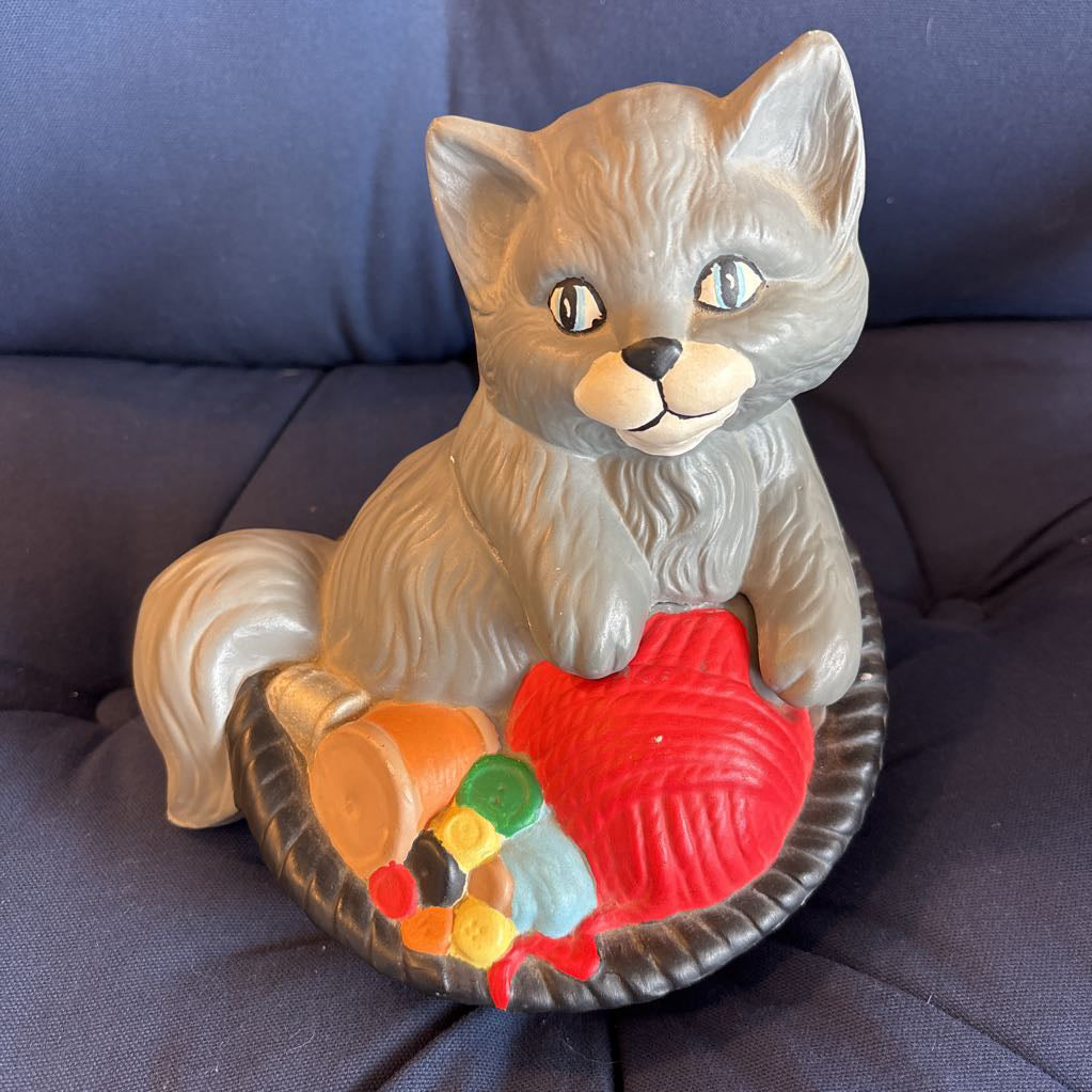 A ceramic figure of a gray cat in a bowl filled with yarn and sewing supplies sitting on a plush navy cushion. 