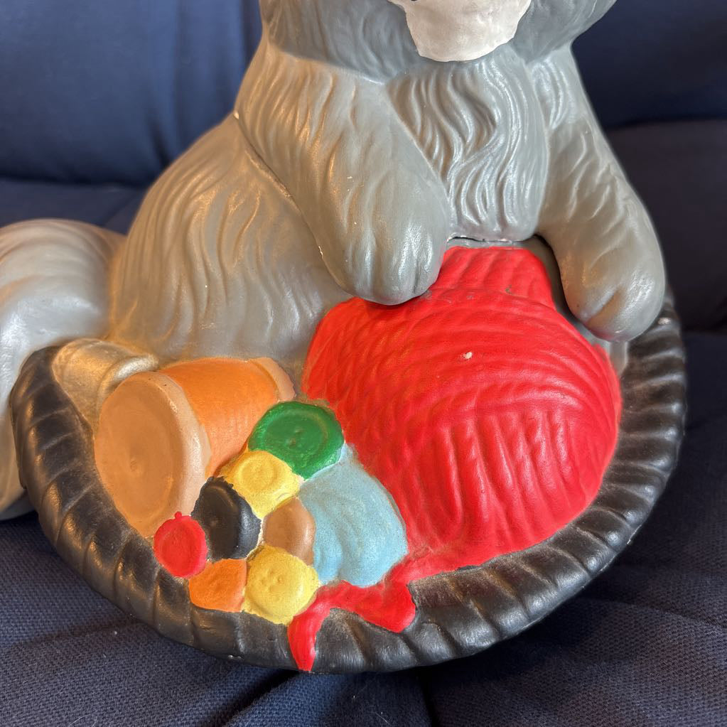 A ceramic figure of a gray cat in a bowl filled with yarn and sewing supplies sitting on a plush navy cushion. 