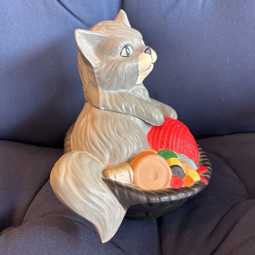 A vintage ceramic cat figurine sitting in a black bowl filled with colorful candy-like pieces, displayed on a dark blue fabric surface.