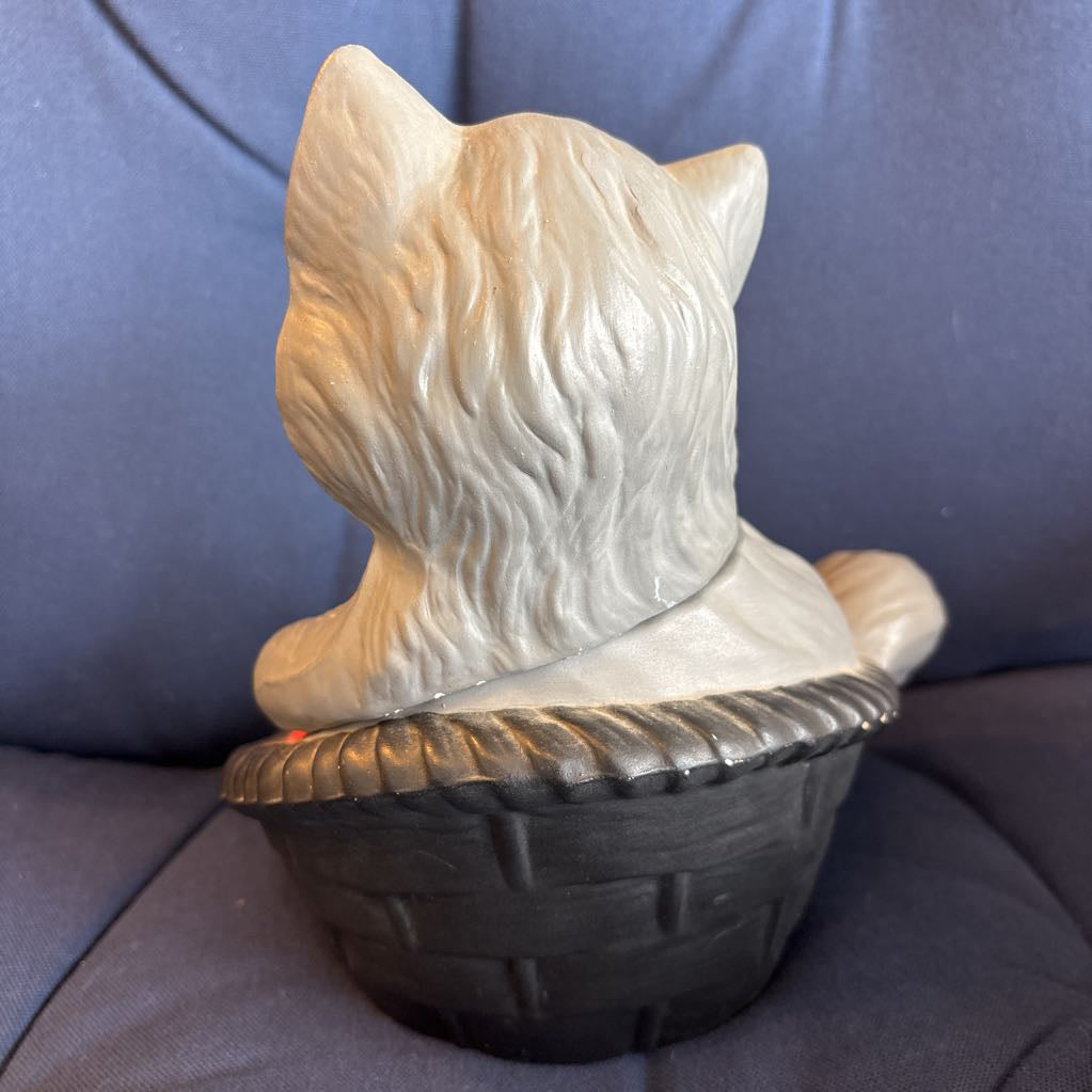 A ceramic cat figurine sitting in a basket, photographed against a blue fabric background.